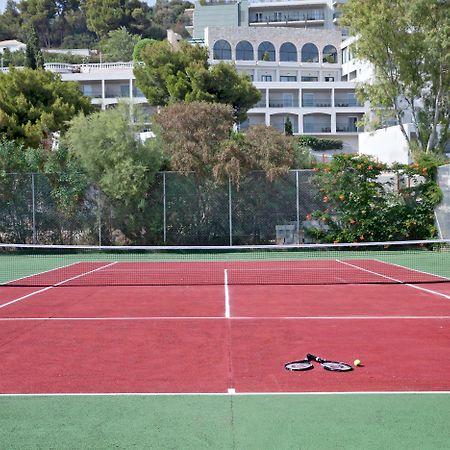ドルチェ バイ ウインダム アッティカ リビエラ ヴラヴローナ エクステリア 写真 The photo shows a tennis court with red and green surfaces. The court is empty, with no players present. In the center, there are a tennis racket and a ball lying on the ground. In the background, there are trees and a building, suggesting the court 