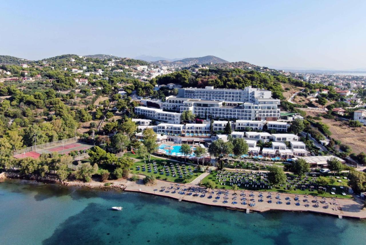ドルチェ バイ ウインダム アッティカ リビエラ ヴラヴローナ エクステリア 写真 The image shows a large hotel complex situated near a beach. It's located on a hillside, surrounded by greenery and trees. The hotel features multiple levels with modern architecture, balconies, and a swimming pool area. In front of the hotel, there'