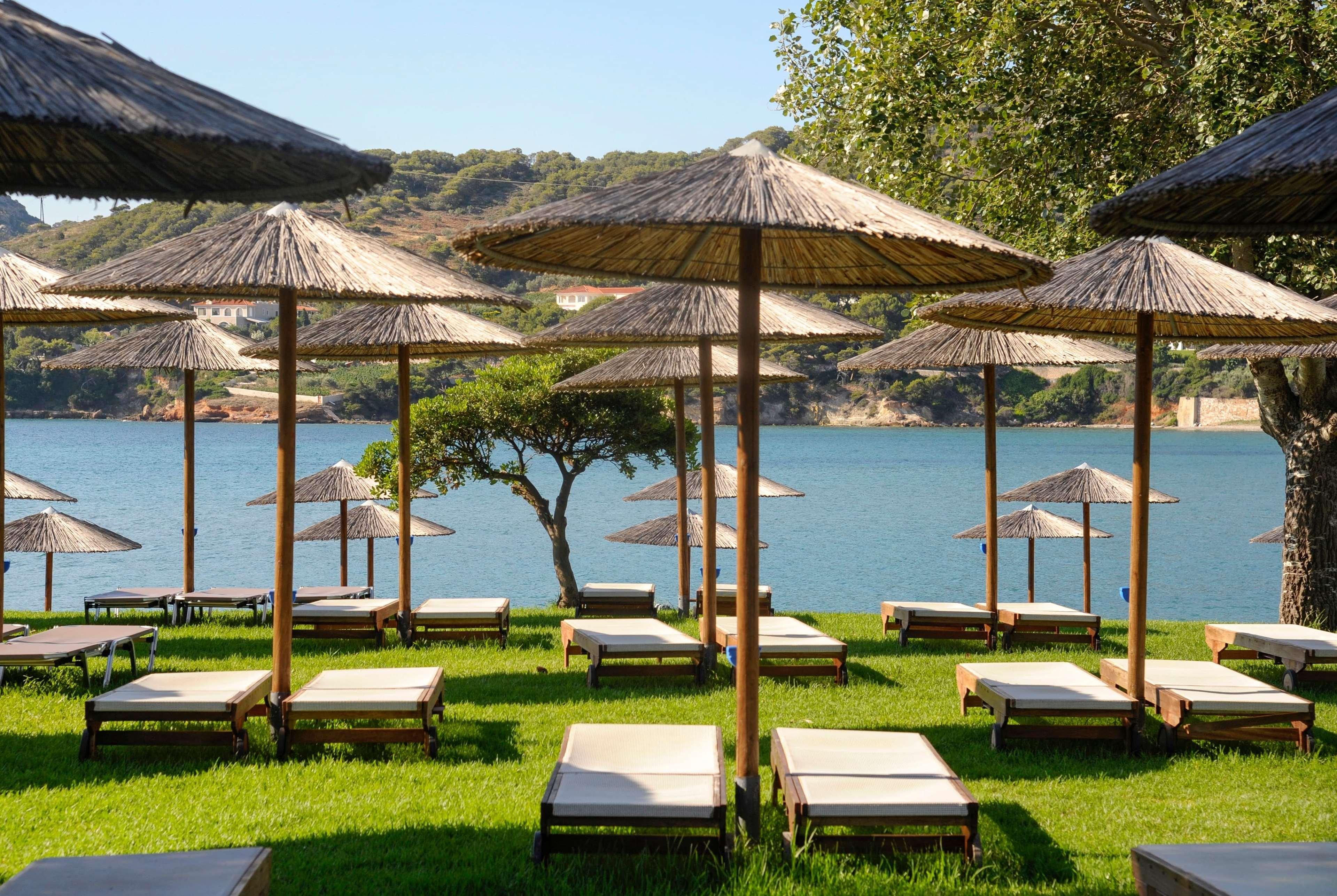 ドルチェ バイ ウインダム アッティカ リビエラ ヴラヴローナ エクステリア 写真 The photo shows a serene beach scene with several lounge chairs arranged on a green lawn. Each lounge chair is shaded by a traditional straw umbrella, providing a relaxing spot to enjoy the view. In the background, there is a calm body of water, like