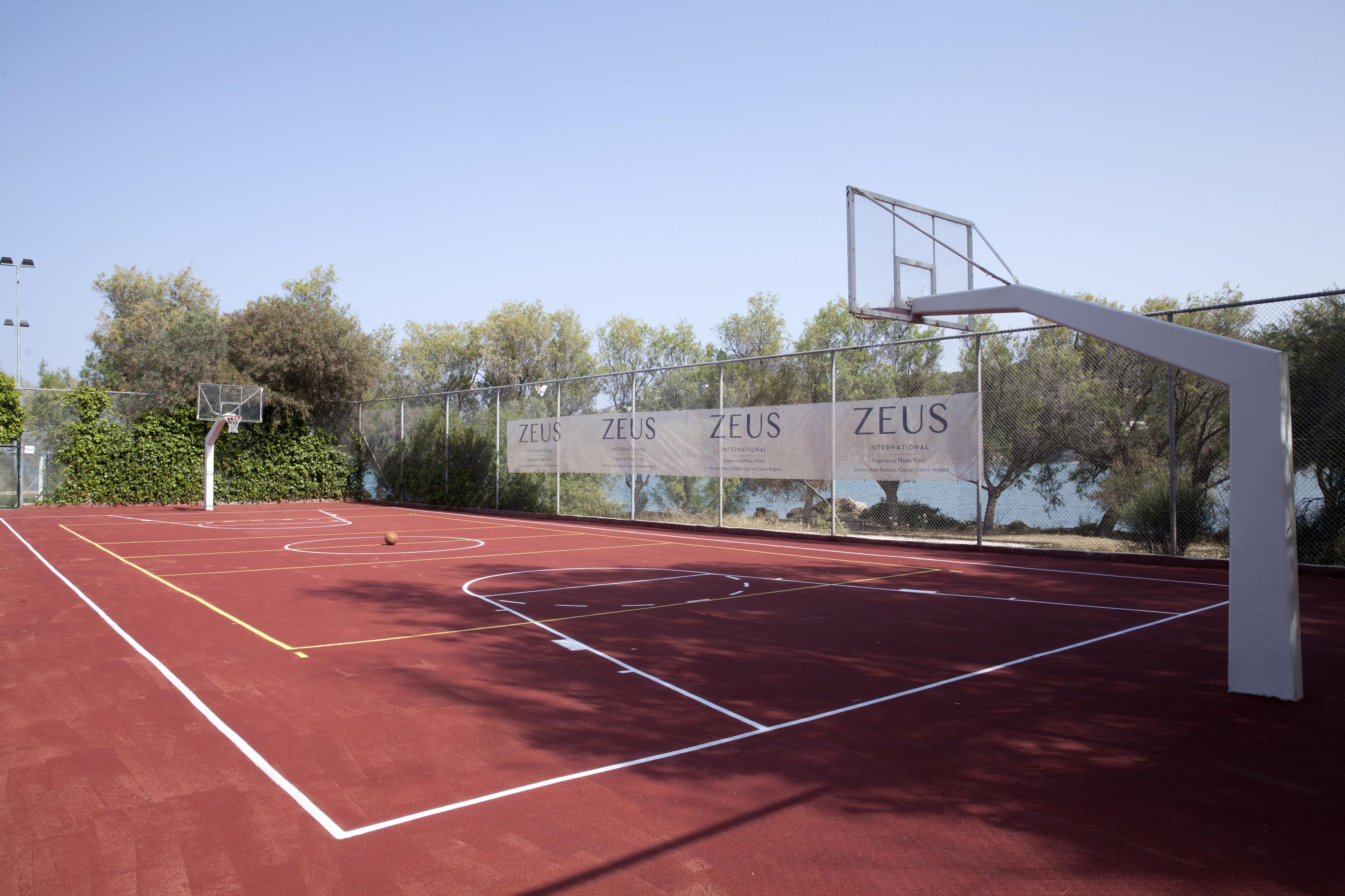 ドルチェ バイ ウインダム アッティカ リビエラ ヴラヴローナ エクステリア 写真 The photo shows a basketball court with a reddish surface. There are two basketball hoops on either side of the court, surrounded by a fence that has banners displaying the word "ZEUS." In the background, there are trees, and the court appears to be 