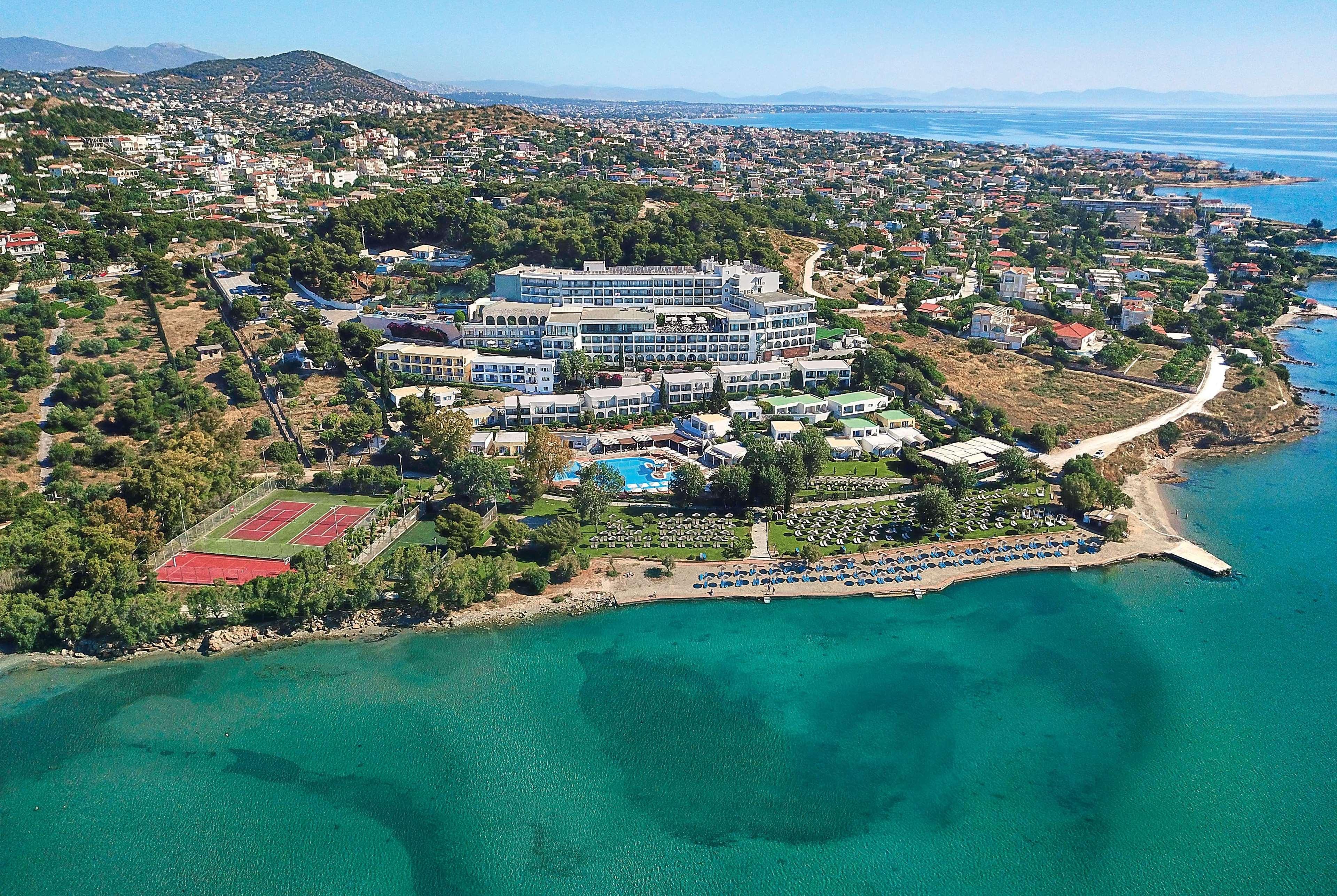 ドルチェ バイ ウインダム アッティカ リビエラ ヴラヴローナ エクステリア 写真 The photo shows an aerial view of a coastal resort area. The resort features multiple buildings, including hotel accommodations and amenities, situated on a hillside overlooking a calm, turquoise sea. There is a sandy shoreline with a few boats on th