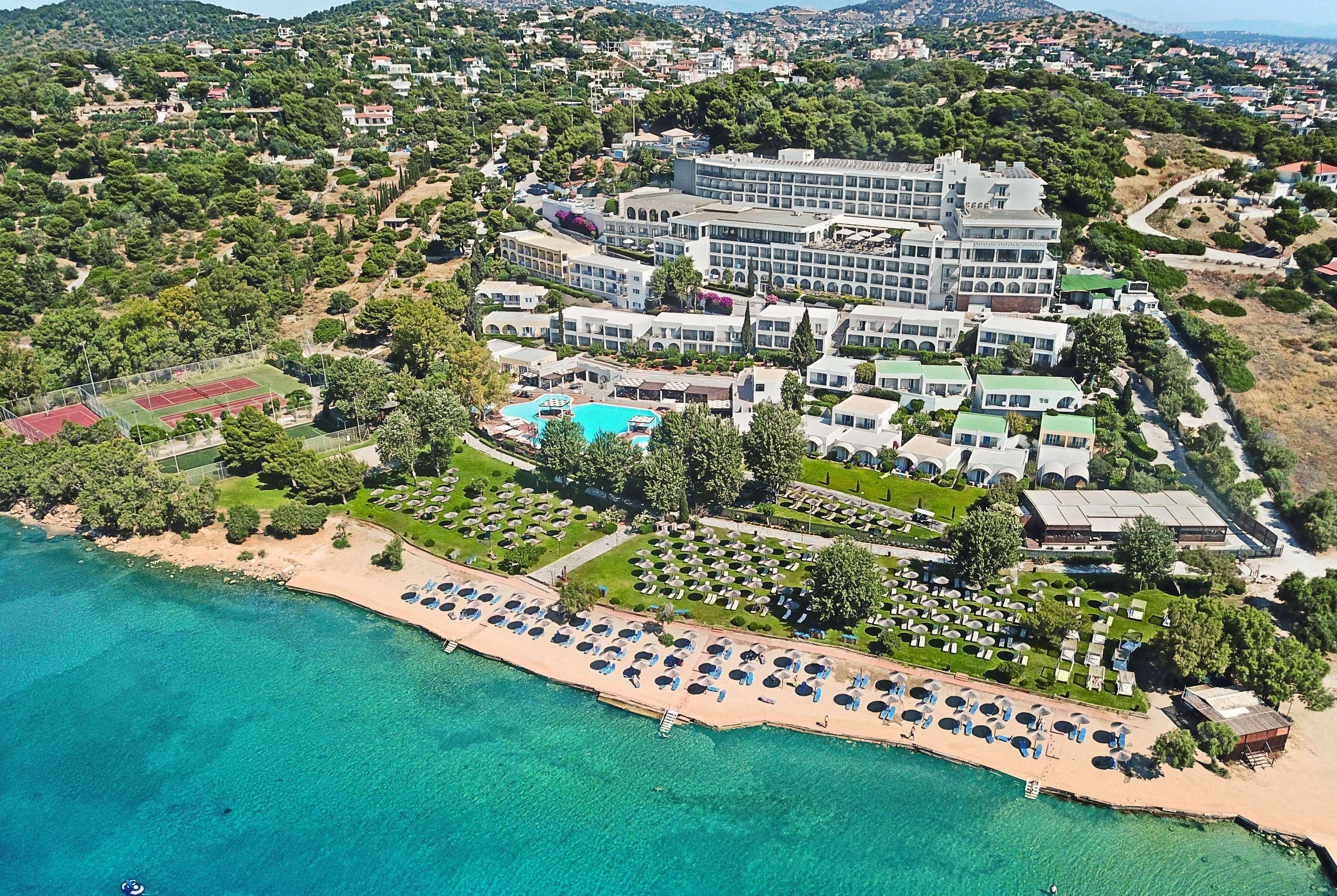 ドルチェ バイ ウインダム アッティカ リビエラ ヴラヴローナ エクステリア 写真 The photo shows a scenic aerial view of a resort located near a beautiful coastline. The resort features a large building and various smaller accommodations. An inviting swimming pool is visible at the center, surrounded by sun loungers and greenery.