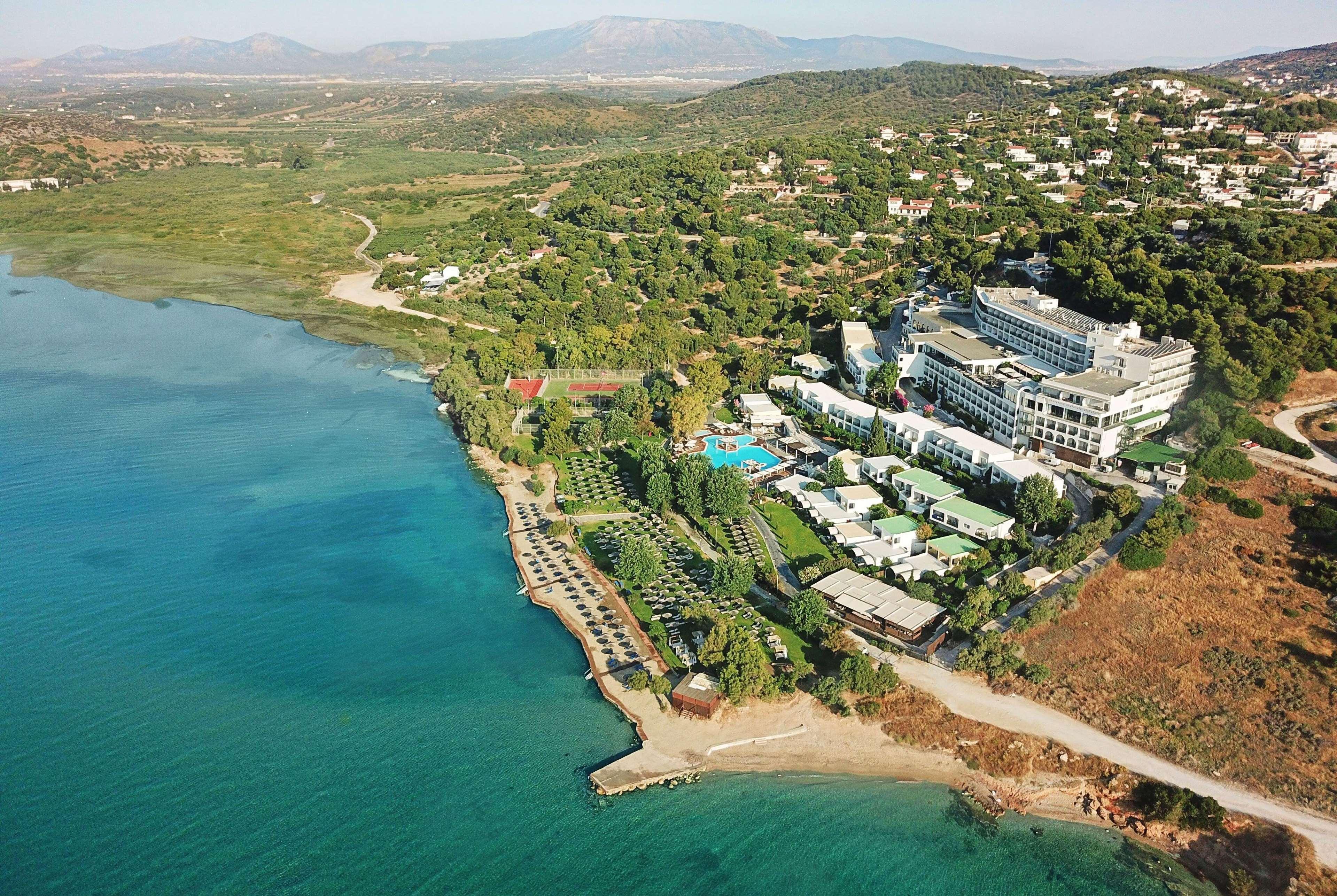 ドルチェ バイ ウインダム アッティカ リビエラ ヴラヴローナ エクステリア 写真 The photo shows an aerial view of a coastal area featuring a resort or hotel complex situated near the shoreline. The building has multiple levels and is surrounded by lush greenery and landscaped gardens. There is a pool area visible, as well as sev