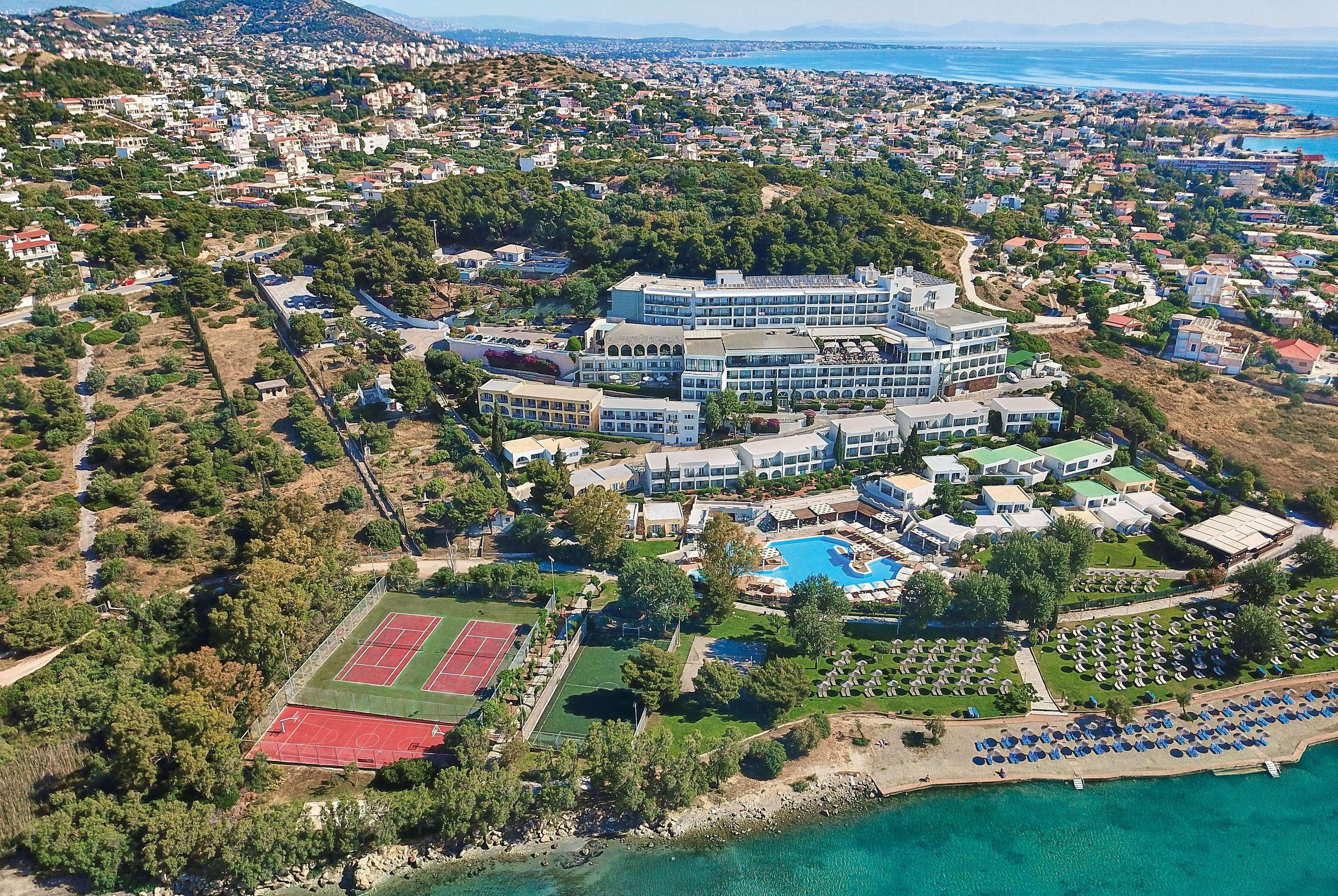 ドルチェ バイ ウインダム アッティカ リビエラ ヴラヴローナ エクステリア 写真 The photo shows an aerial view of a resort or hotel complex situated near the coast. The building appears to be large, with multiple levels and several balconies. Surrounding the hotel are green landscaped areas, including gardens and perhaps a pool.