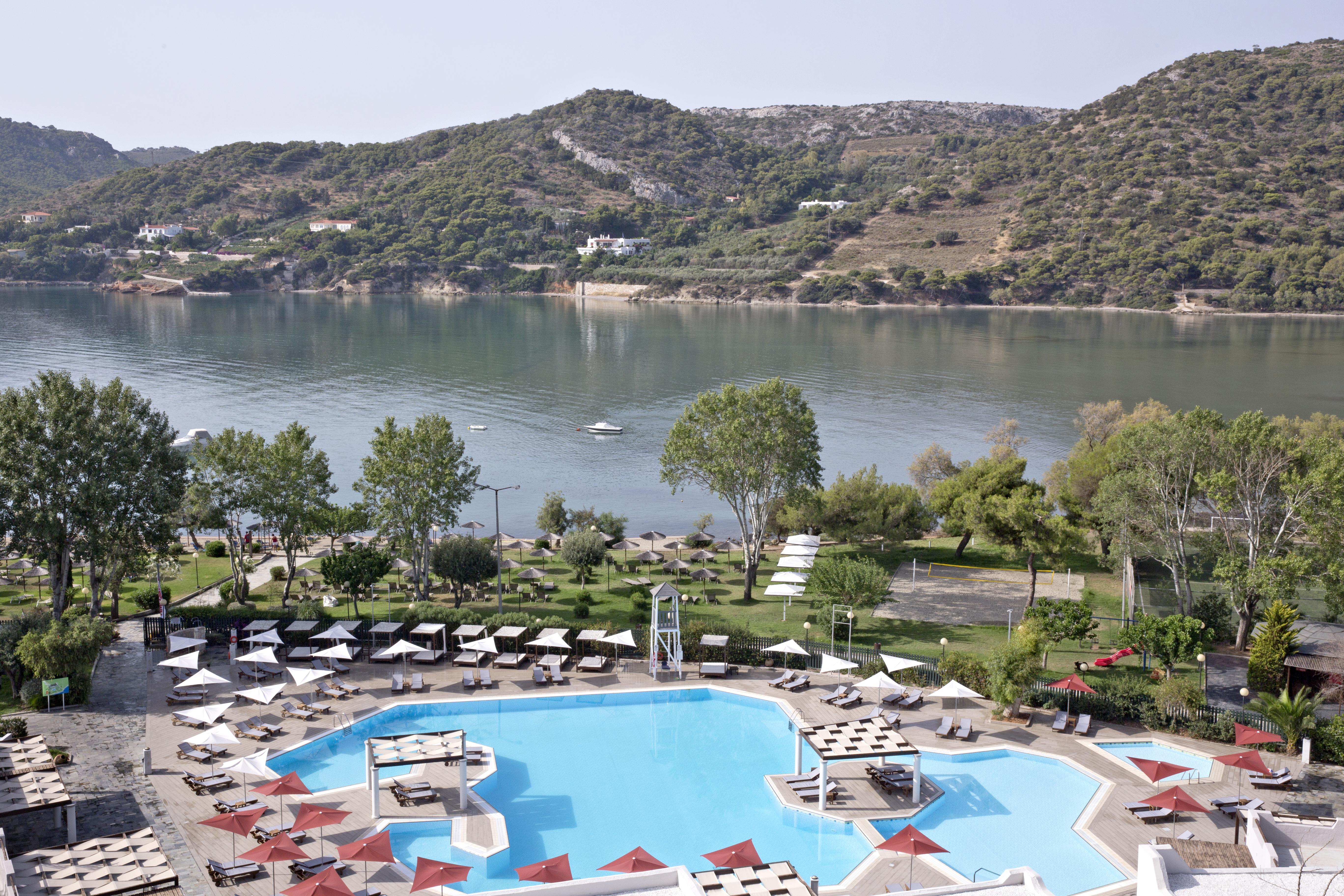 ドルチェ バイ ウインダム アッティカ リビエラ ヴラヴローナ エクステリア 写真 The photo depicts a serene view of a resort area featuring a large swimming pool in the foreground. The pool has a unique octagonal shape and is surrounded by sun loungers and umbrellas, creating a relaxing atmosphere. Beyond the pool, there is a cal
