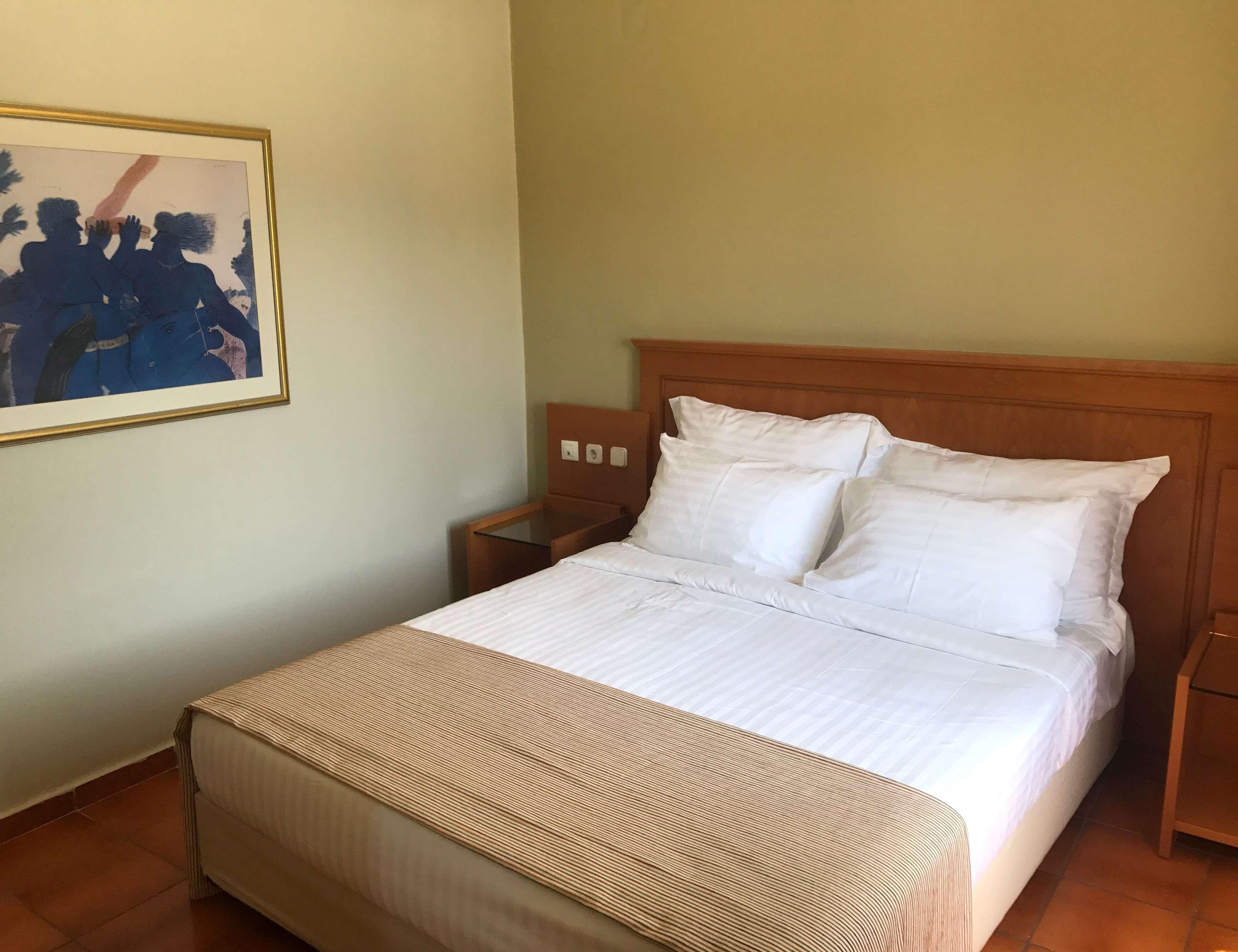 ドルチェ バイ ウインダム アッティカ リビエラ ヴラヴローナ エクステリア 写真 The photo shows a simple and clean hotel room. It features a double bed with white linens and two pillows, and a beige bed covering. On the left side, there is a framed painting or wall art hung on the wall. The room has light-colored walls and woode