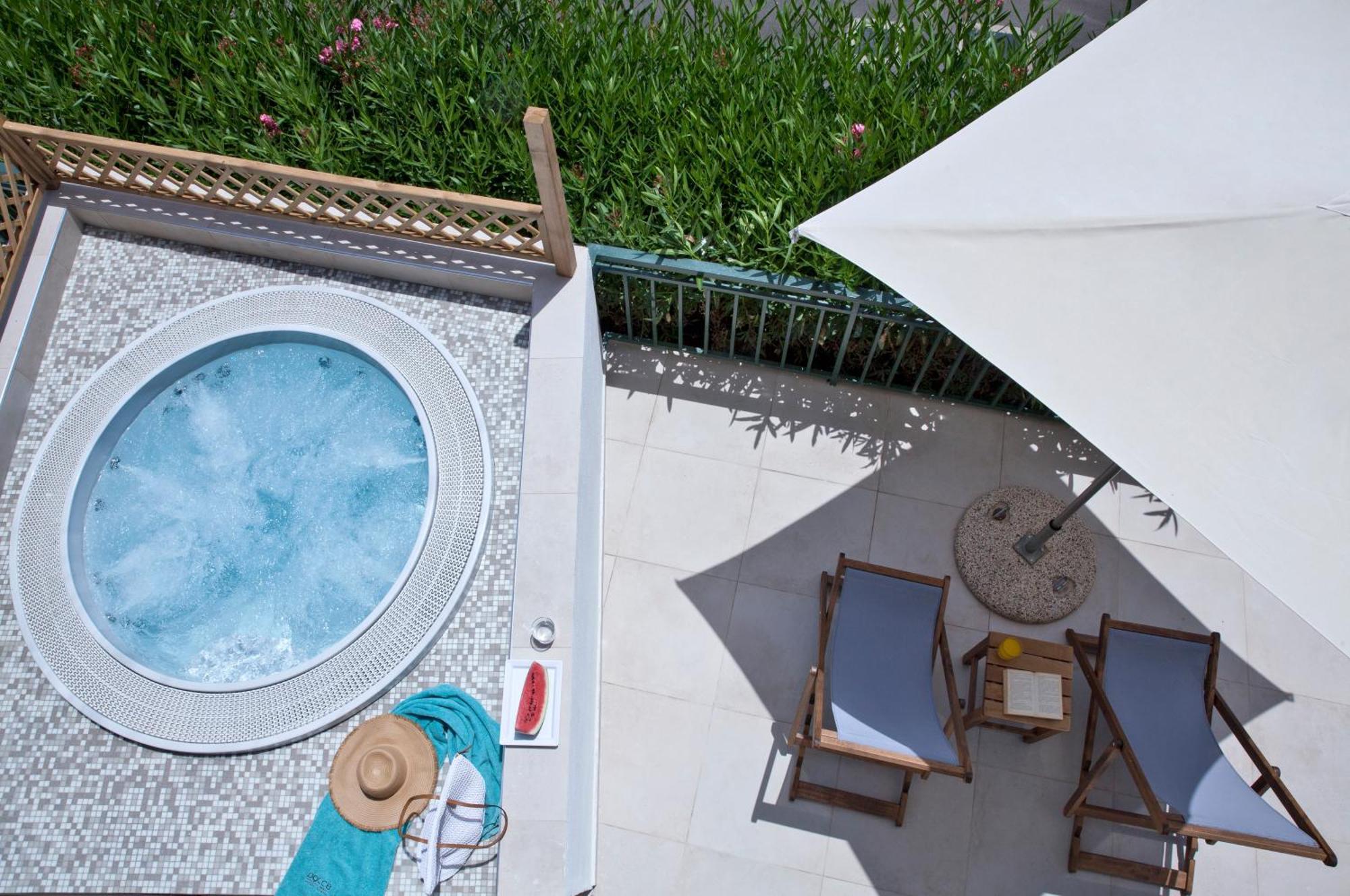 ドルチェ バイ ウインダム アッティカ リビエラ ヴラヴローナ エクステリア 写真 The photo shows an overhead view of a relaxing outdoor space. There's a round hot tub with light blue water and a decorative edge. Nearby, a couple of wooden lounge chairs are positioned underneath a large umbrella to provide shade. A small side tabl