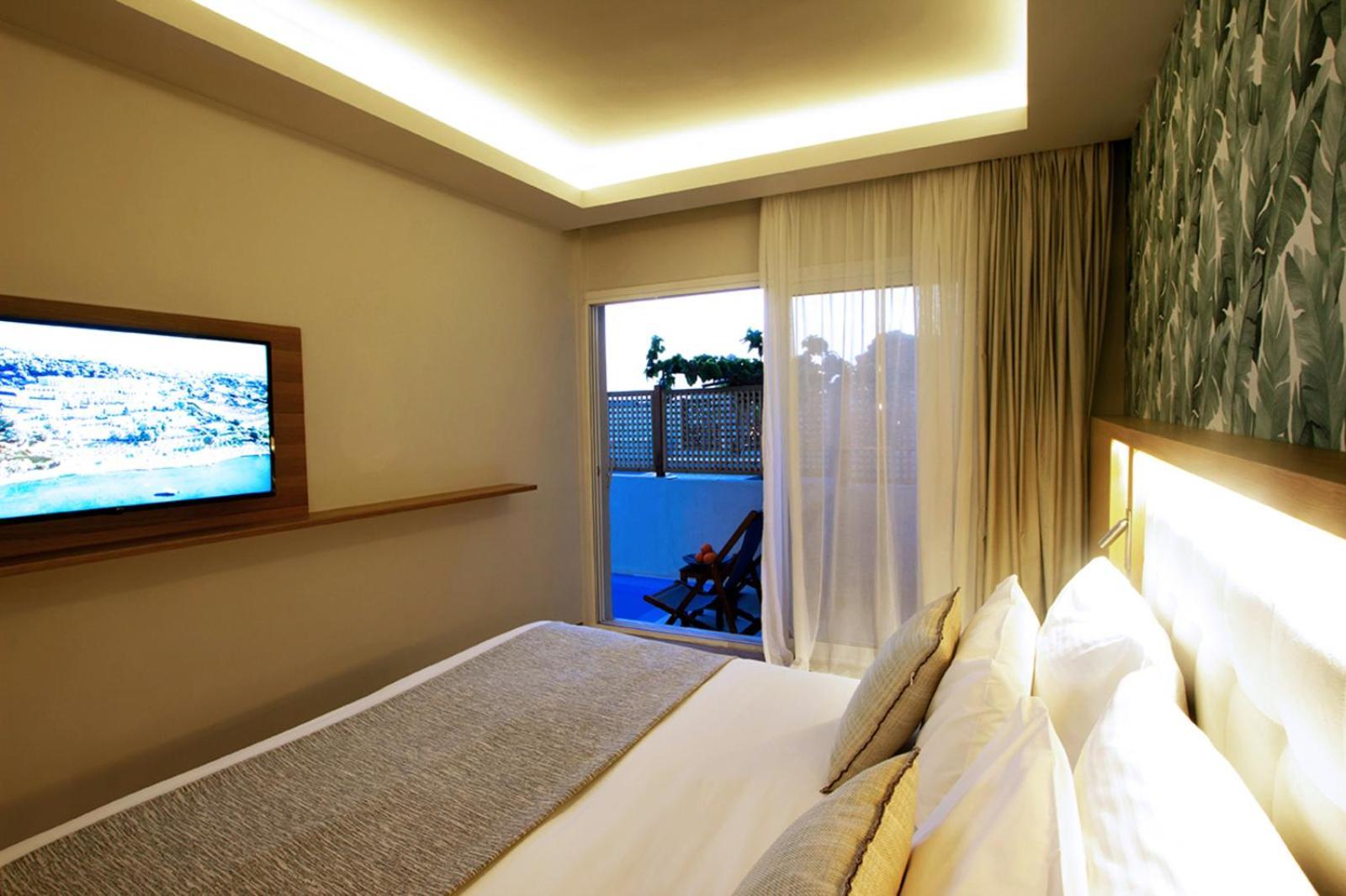 ドルチェ バイ ウインダム アッティカ リビエラ ヴラヴローナ エクステリア 写真 The photo shows a modern hotel room featuring a large bed with white linens and decorative pillows. There's a mounted flat-screen television on the wall opposite the bed. Natural light enters the room through sheer curtains, leading to a balcony or o